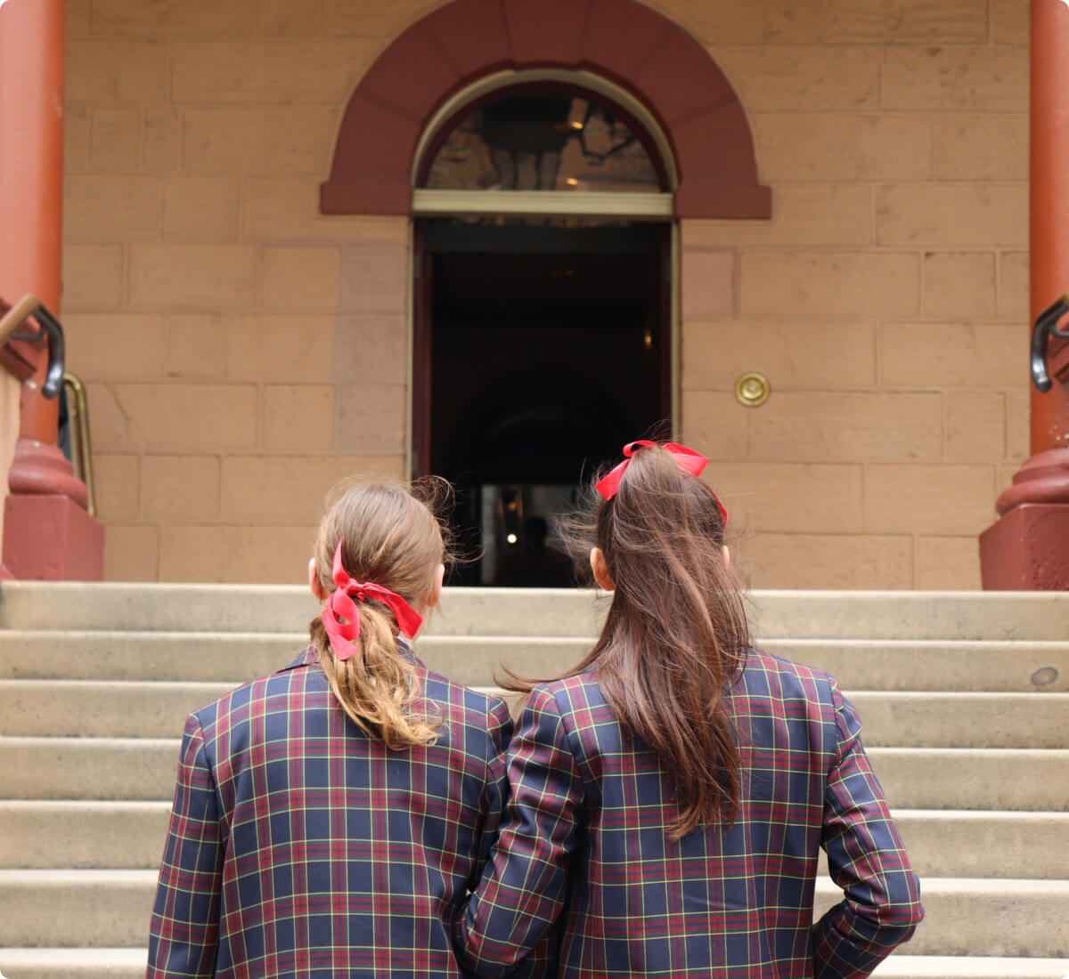 Two girls with red hair ribbons and plaid uniforms stand together, back-facing, at the base of stone steps leading to an arched building entrance.